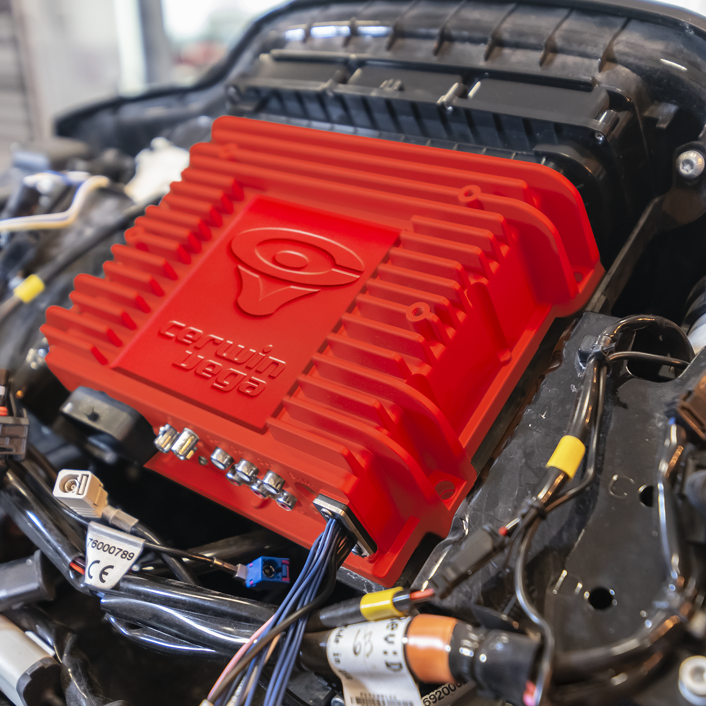 A close-up of an engine shows a red component with the Cerwin Vega logo, reminiscent of Harley Davidson style. This ribbed part, surrounded by black and metallic wires and connectors, is the 2014-2023 Harley Davidson® Plug N-Play Media Receiver - RPMHD14.