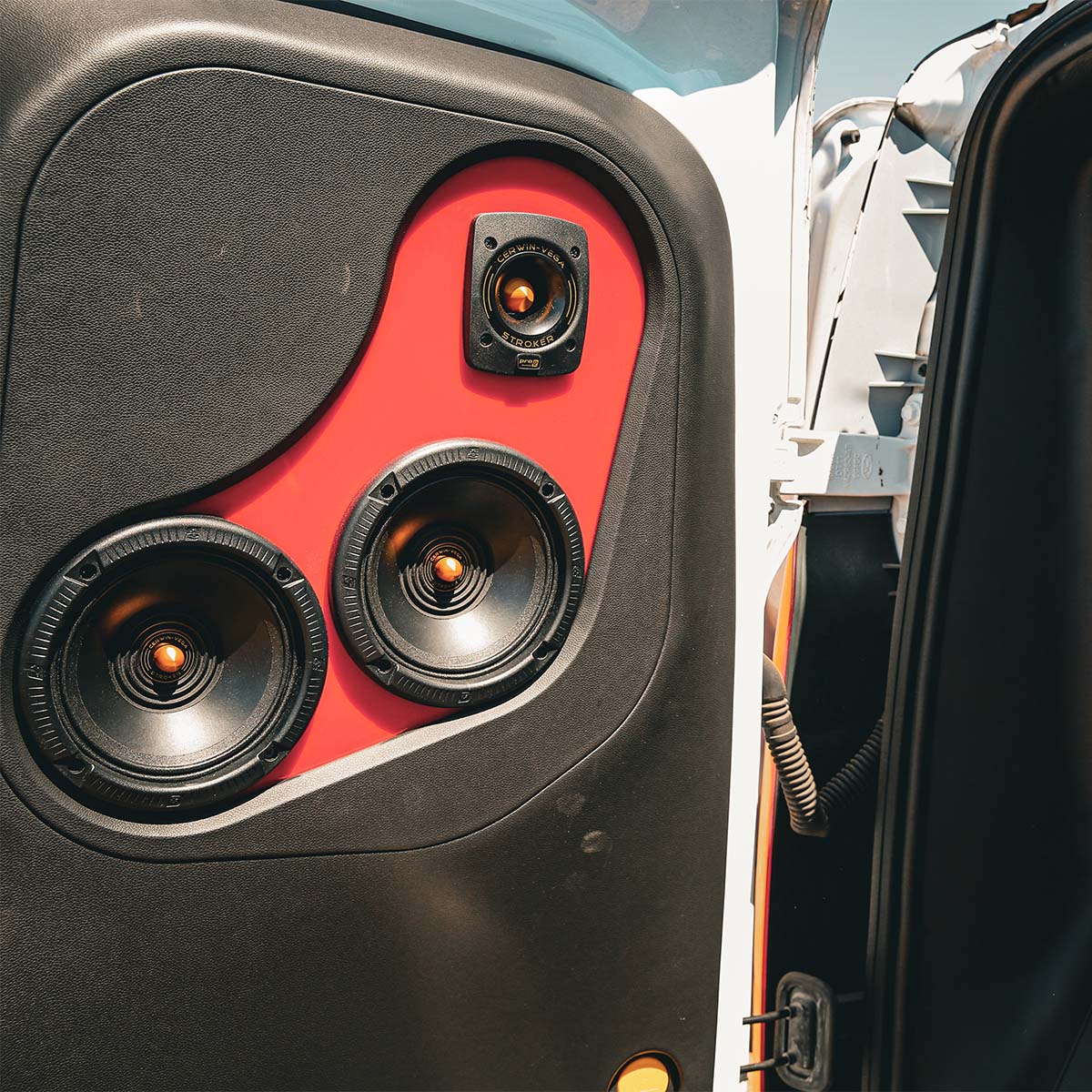 Close-up of a car door panel featuring Cerwin Vega's Stroker Pro CVMPCL6.5 audio system against a red background, showcasing three circular speakers for exceptional clarity, with the largest at the bottom. The open door reveals part of the car's interior and a blue sky outside.