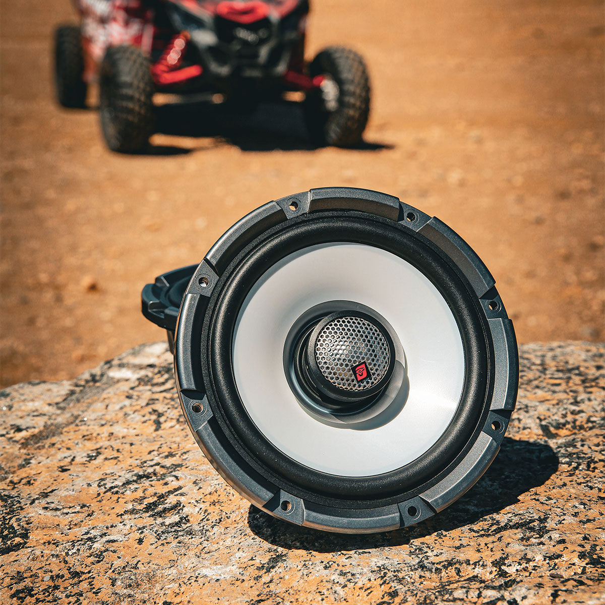 A close-up of the RPM Stroker SM65F4, a black and white round 6.5" car speaker featuring a flush mount 1" titanium dome tweeter, is set on textured stone. In the blurred background, a red and black remote-controlled car appears on sandy terrain, highlighting an outdoor tech ambiance.