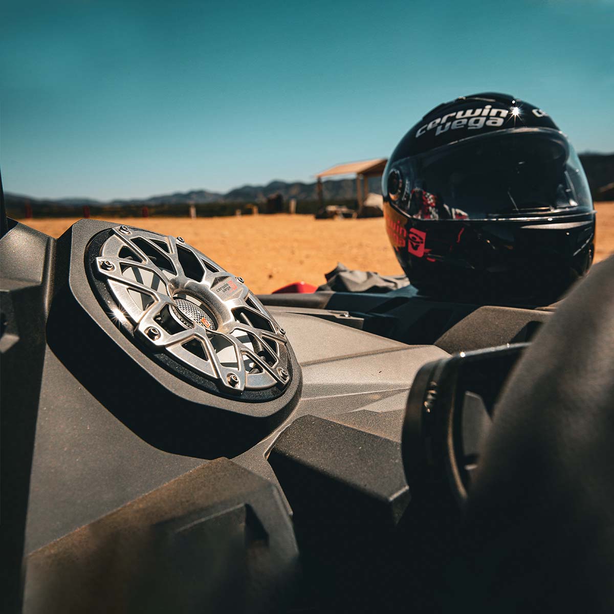 Close-up of a vehicle dashboard highlighting the RPM Stroker SM65F4 2-Way 6.5" Speaker with flush mount titanium dome tweeter and a black helmet. The sunny, sandy landscape with distant mountains and a structure under clear blue skies in the background suggests an enticing adventure setting.