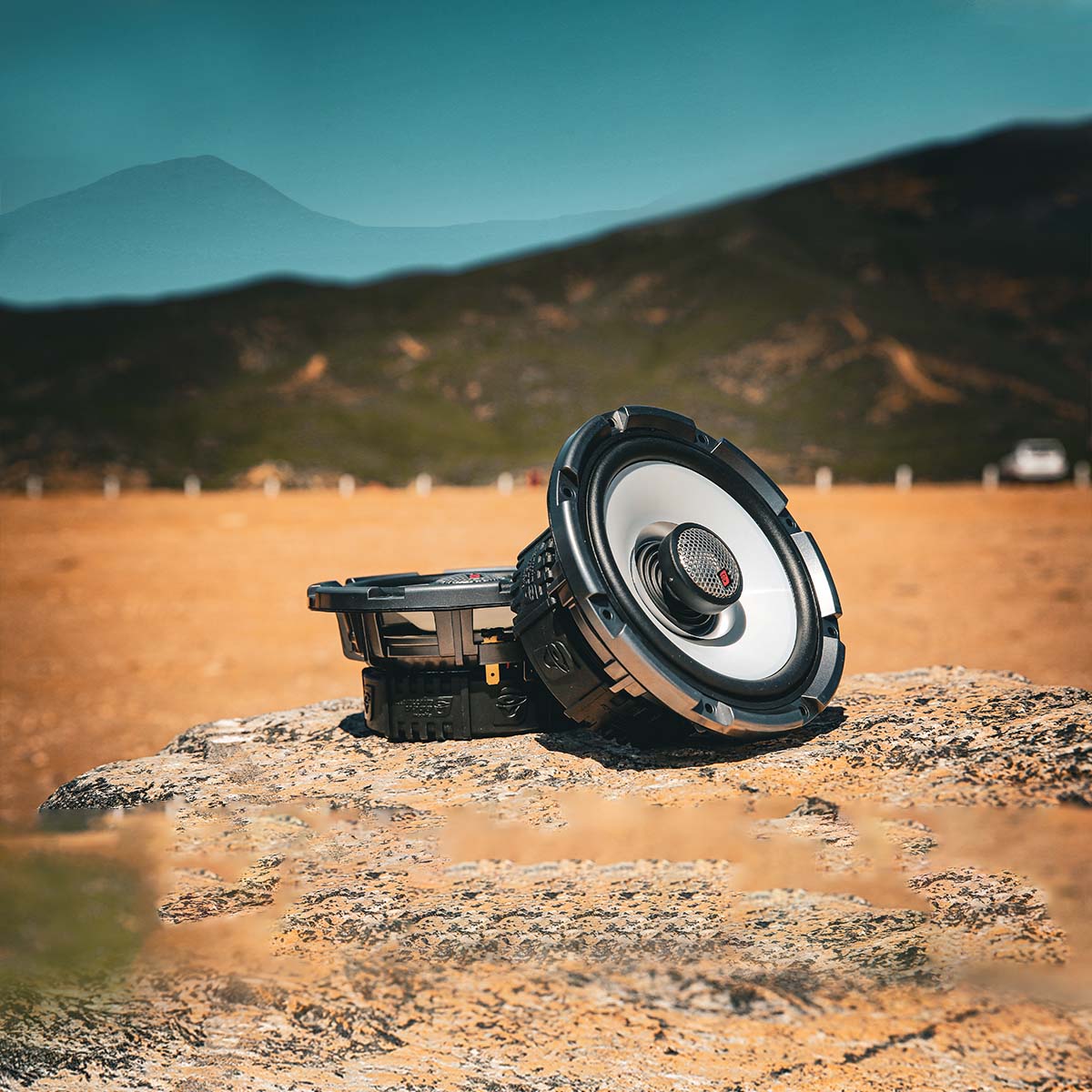 The RPM Stroker SM65F4, a sleek 2-way 6.5" speaker with a flush mount 1" titanium dome tweeter, sits weather-resistant on a rugged rock in the desert. Its modern design features a prominent central cone and dark casing under a clear blue sky against towering mountains, suggesting a sunny day.
