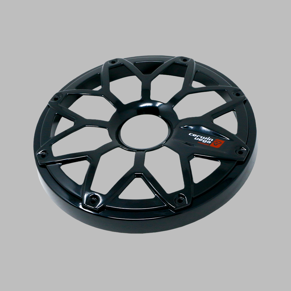 A black circular bike wheel cover with an intricate geometric cutout design resembles an RPM Stroker 6.5" Flush Mount Speaker Grill. The text "Corima 50th" appears in white and red on a small black panel near the edge of the plain gray background.