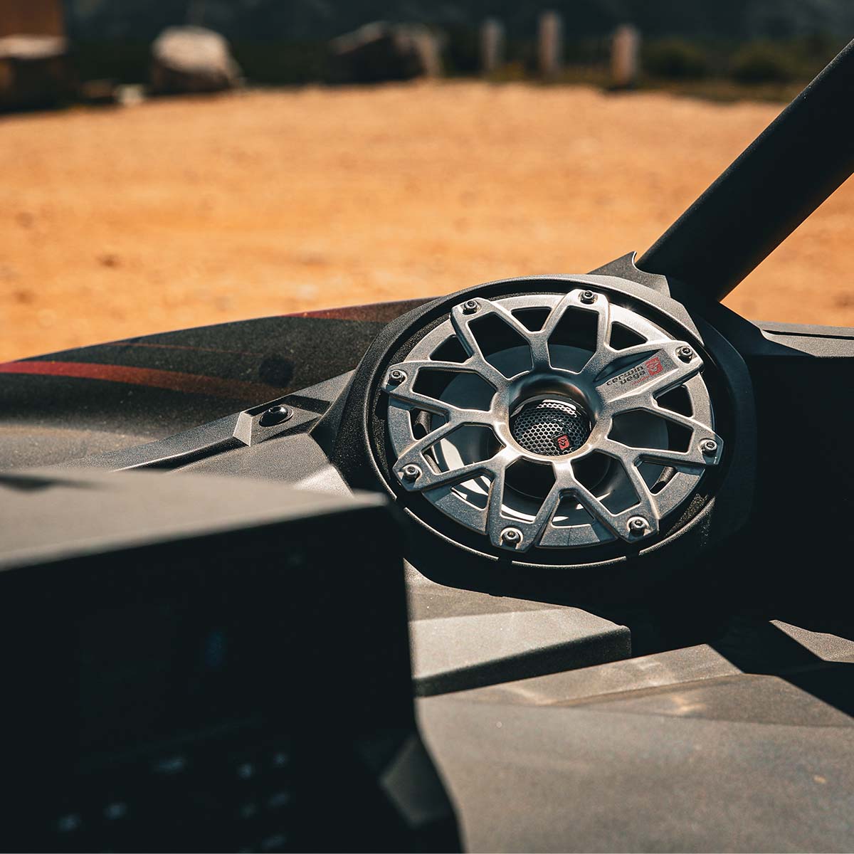 A close-up showcases the RPM Stroker 8" Flush Mount Speaker Grill with a metallic geometric design. Set against a sandy landscape, sunlight highlights its intricate details, creating a rugged ambiance with shadows on the grill.