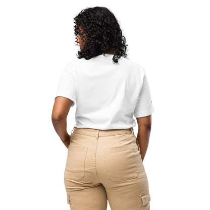 A person with curly hair, seen from the back, wears a Cerwin-Vega Breast Cancer Awareness T-Shirt, a white short-sleeved shirt featuring a small pink ribbon. Beige pants with pockets complete the casual look against the plain white background.