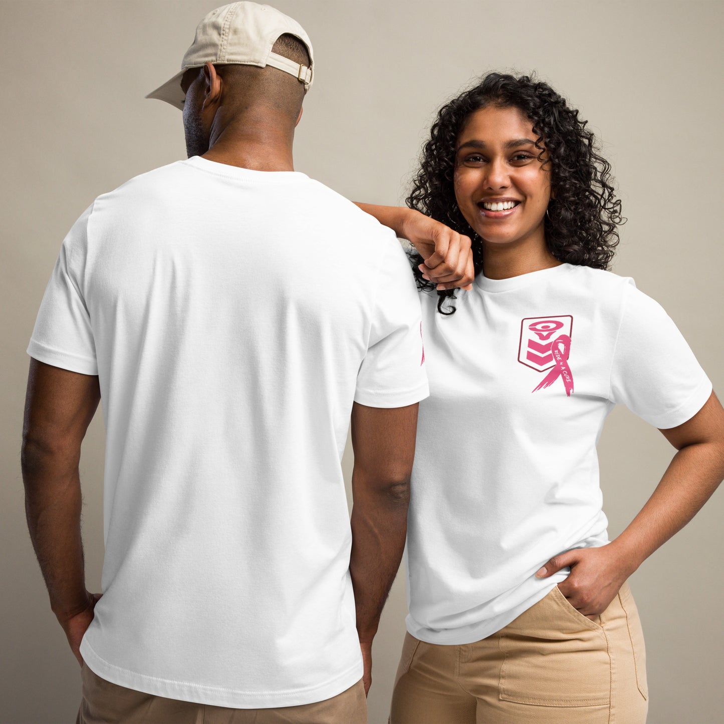 A smiling woman with curly hair wears a Cerwin-Vega Breast Cancer Awareness T-shirt featuring a pink ribbon. Beside her, a man showcases the back of the same white T-shirt and wears a beige cap. Both are dressed in beige pants against a neutral background.